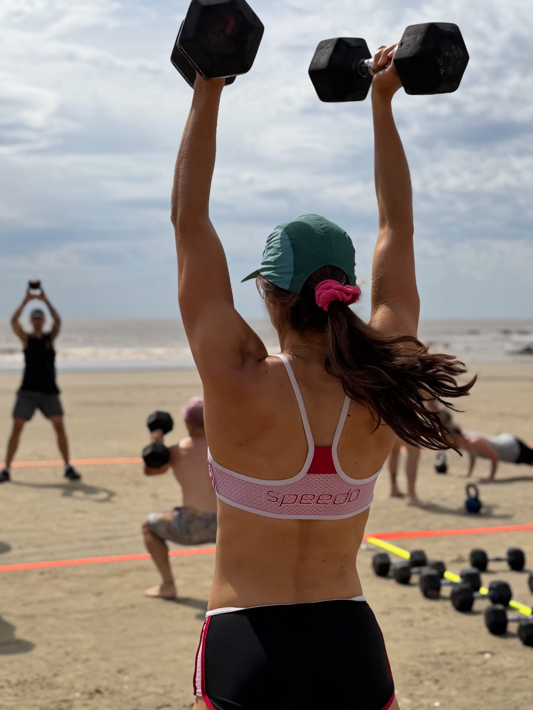 Camila pressing dumbbells overhead during a beach workout